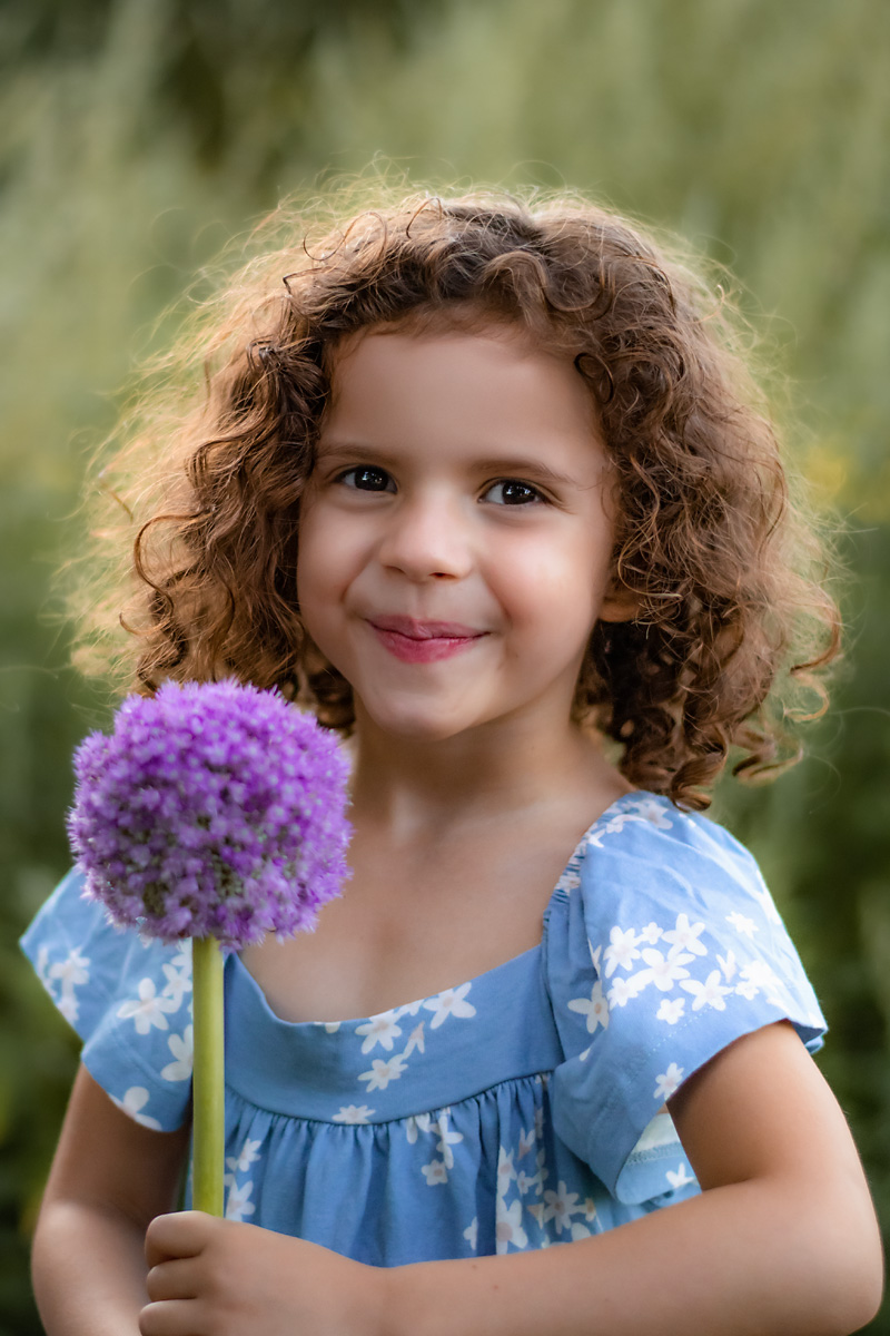Ein kleines Mädchen mit lockigem Haar lächelt in die Kamera und hält eine große lila Blume bei einem Kinderfotoshooting im Freien in Deutschland.