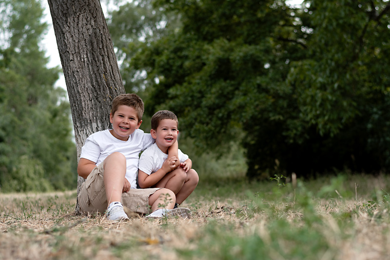 Zwei lachende Brüder in weißen T-Shirts sitzen zusammen unter einem Baum auf der Wiese bei einer Kindergartenfotografie im Freien in Deutschland.