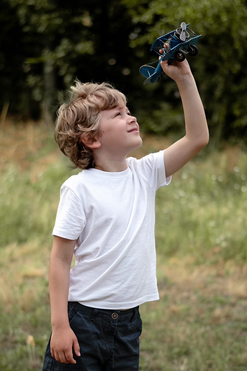 Ein kleiner Junge im weißen T-Shirt hält ein blaues Spielzeugflugzeug hoch in die Luft bei einer Kindergartenfotografie im Freien in Deutschland.