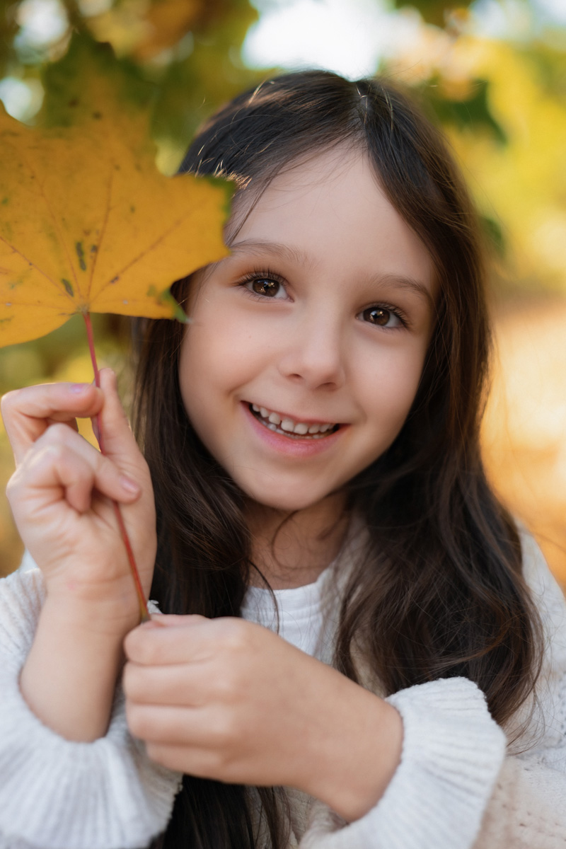 Nahaufnahme eines lächelnden Mädchens mit langen Haaren, das ein gelbes Herbstblatt neben ihr Gesicht hält bei einer Kindergartenfotografie in Deutschland.