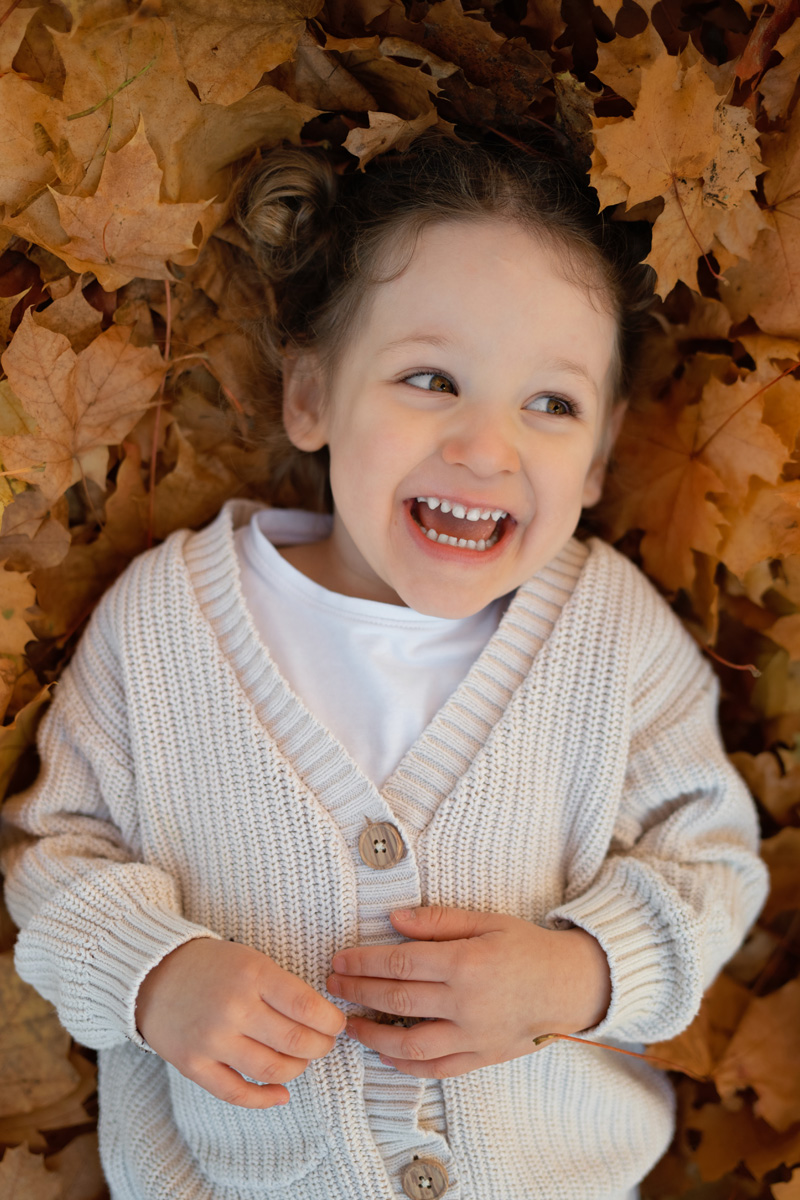 Ein kleines Mädchen liegt fröhlich lachend im bunten Herbstlaub bei einer Kindergartenfotografie im Freien in Deutschland.