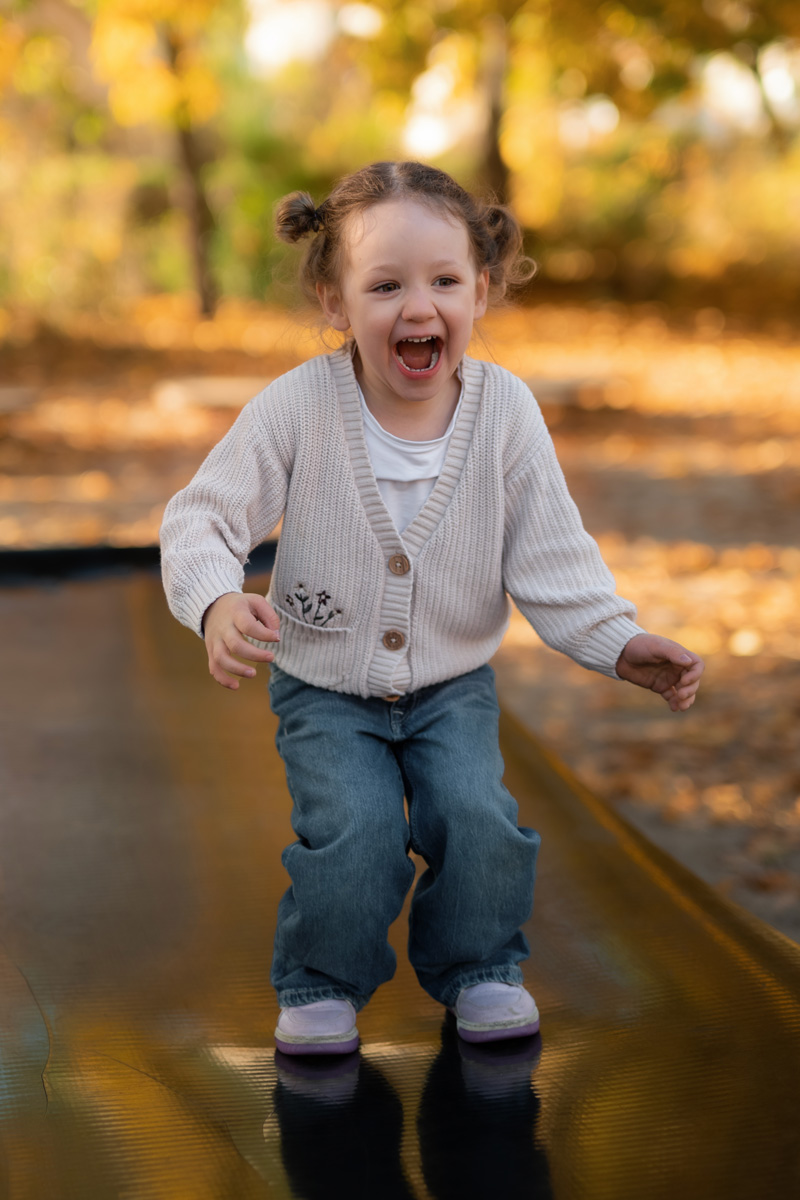Ein kleines Mädchen springt fröhlich und lachend auf einem Hüpfkissen im Herbst bei einer Kindergartenfotografie in Deutschland.