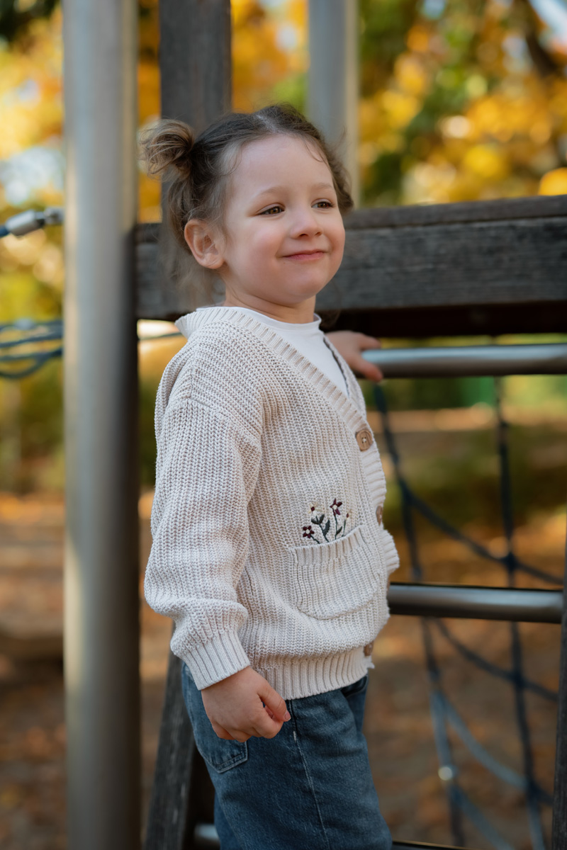 Ein kleines Mädchen im gestrickten Cardigan steht an einem Klettergerüst auf dem Spielplatz und lächelt während einer Kindergartenfotografie in Deutschland.
