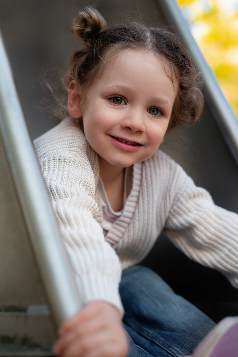 Ein kleines Mädchen mit Hochsteckfrisur lächelt in die Kamera, während es auf einer Rutsche auf dem Spielplatz sitzt bei einer Kindergartenfotografie in Deutschland.