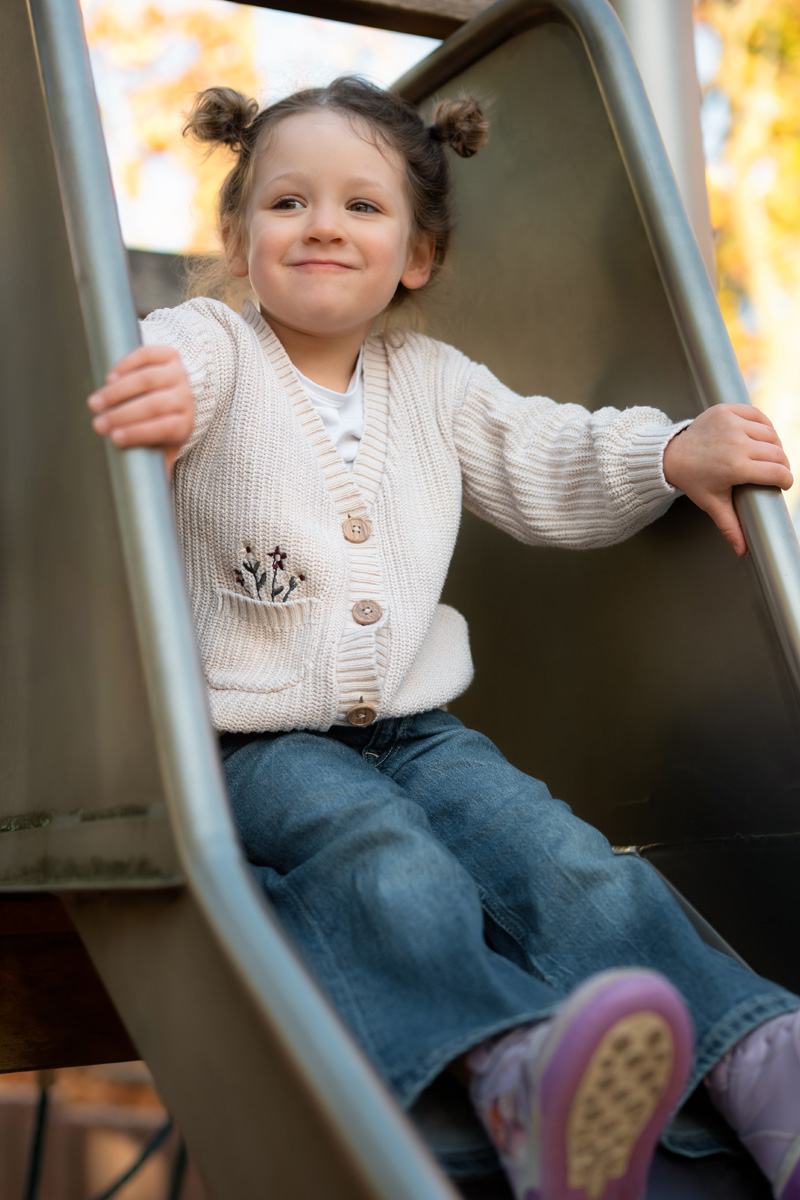 Ein kleines Mädchen mit Zöpfen und hellen Strickjacke lächelt vergnügt, während es auf einer Spielplatzrutsche sitzt bei einer Kindergartenfotografie in Deutschland.