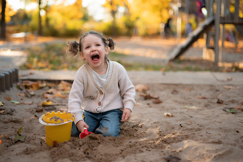 Ein kleines Mädchen mit Zöpfen lacht fröhlich beim Spielen im Sandkasten mit Eimer und Schaufel auf einem Spielplatz bei einer Kindergartenfotografie in Deutschland.