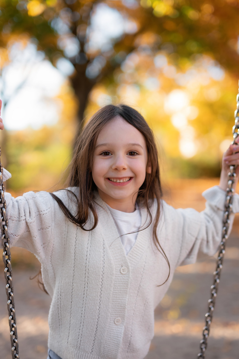 Ein lachendes Mädchen mit langen Haaren sitzt auf einer Schaukel vor herbstlicher Natur bei einer Kindergartenfotografie in Deutschland.