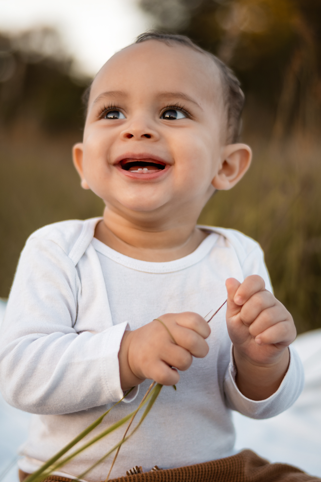 Ein süßes Baby im weißen Body sitzt im Freien auf einer Decke, hält einen Grashalm und lächelt fröhlich nach oben. Natürliche Baby- und Familienfotografie in Deutschland.