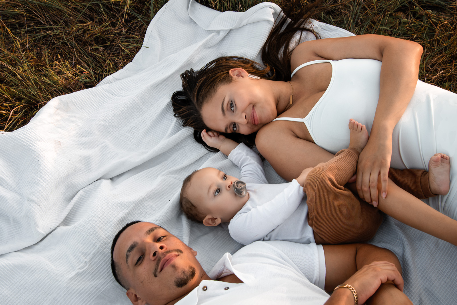 Eine junge Familie liegt gemeinsam mit ihrem Baby auf einer weißen Decke im Gras bei einem Picknick im Freien in Deutschland.