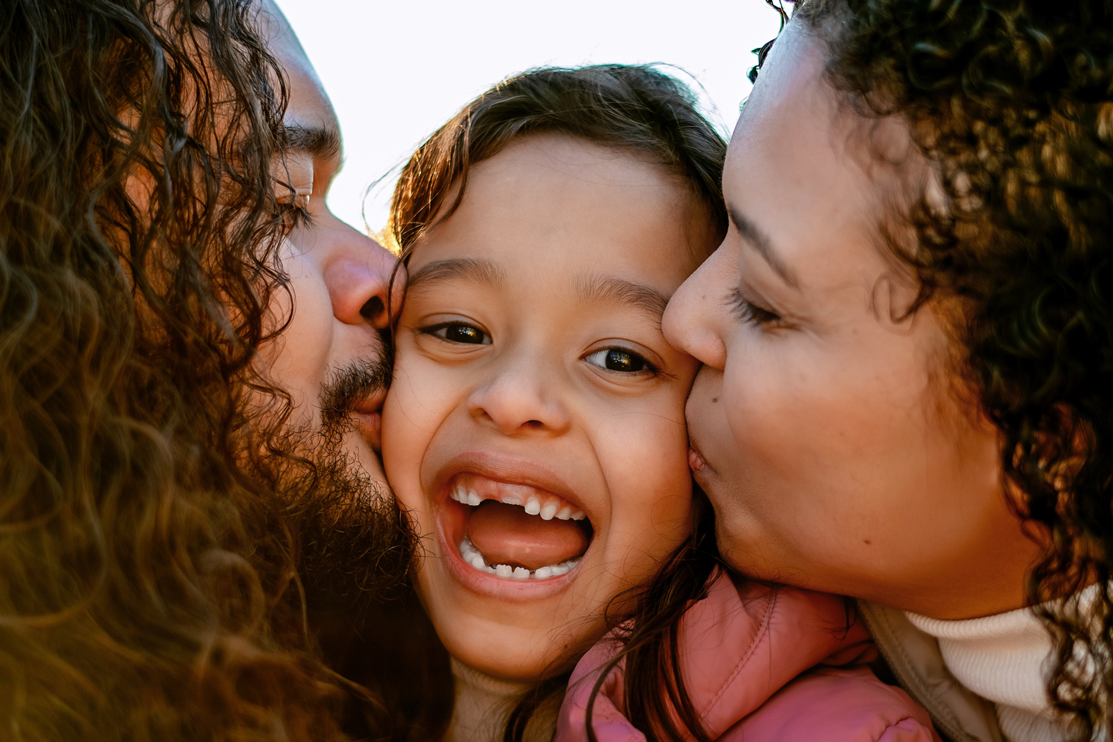Glückliche Eltern küssen ihr lachendes Kind gleichzeitig auf die Wangen bei einem Familien-Fotoshooting im Freien in Deutschland.