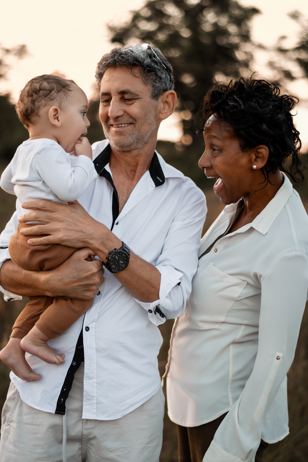 Ein glückliches älteres Paar hält ein kleines Baby im Arm und blickt es freudig an. Emotionale Familienfotografie im Freien in Deutschland.
