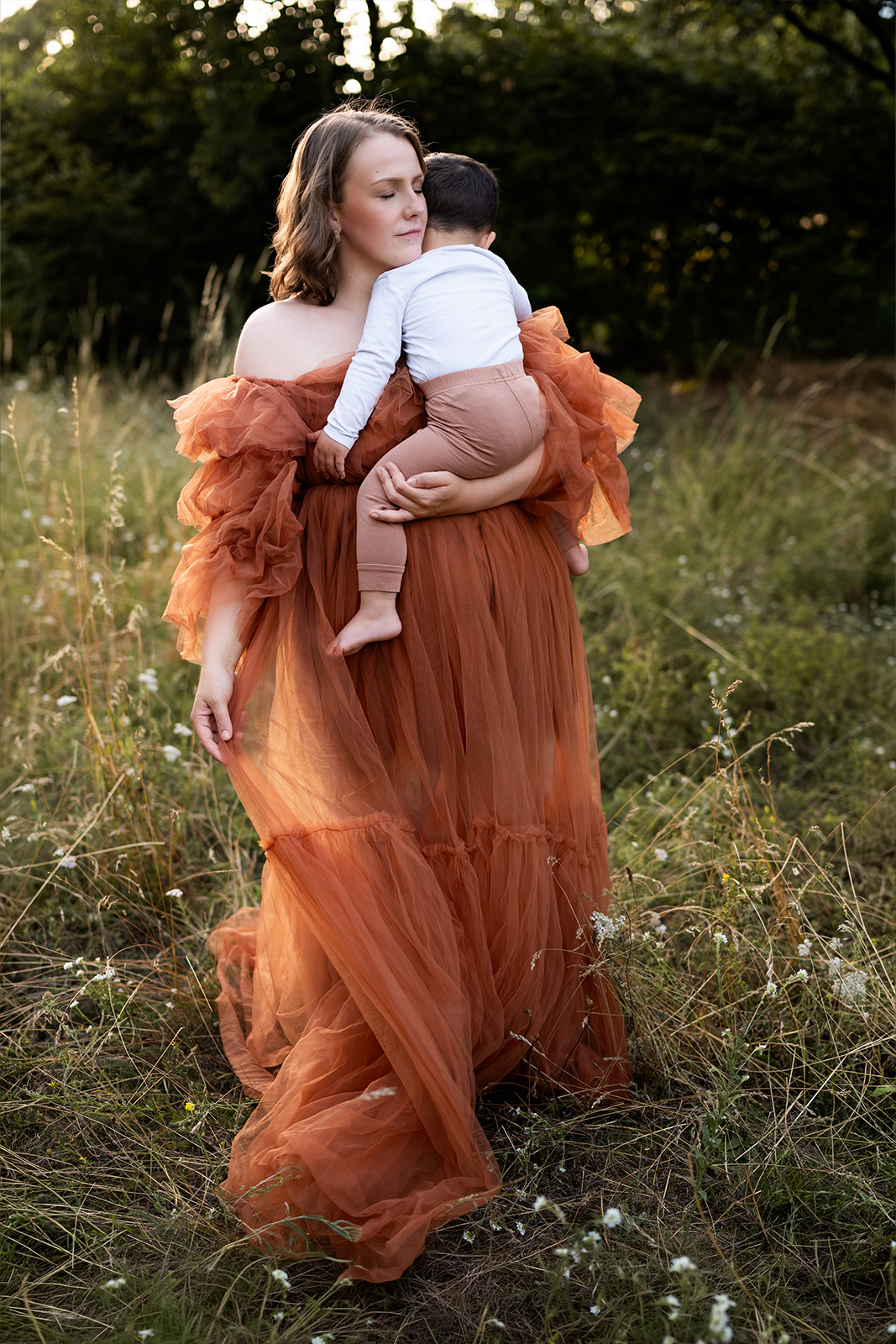 Eine Mutter im eleganten braunen Tüllkleid steht in hohem Gras und hält ihr kleines Kind sanft im Arm. Emotionale Familienfotografie in Deutschland.