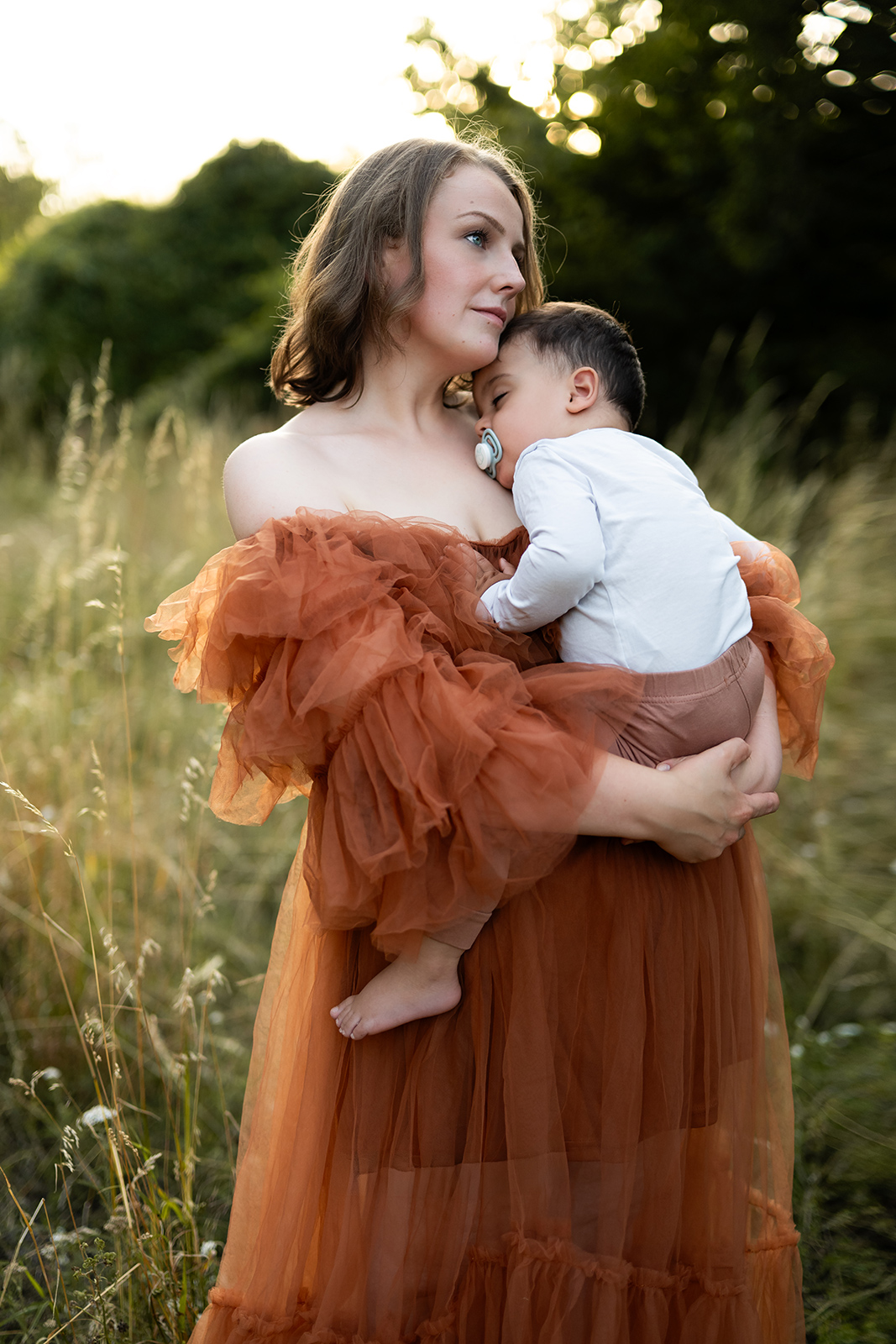 Eine elegante Mutter im braunen Tüllkleid hält ihr schlafendes Baby sanft im Arm auf einer grünen Wiese. Ästhetische Familienfotografie in Deutschland