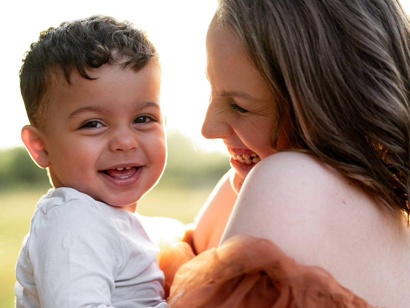 Ein lachendes kleines Kind schaut in die Kamera, während seine Mutter es liebevoll im Arm hält. Natürliche Familienfotografie im Freien in Deutschland.