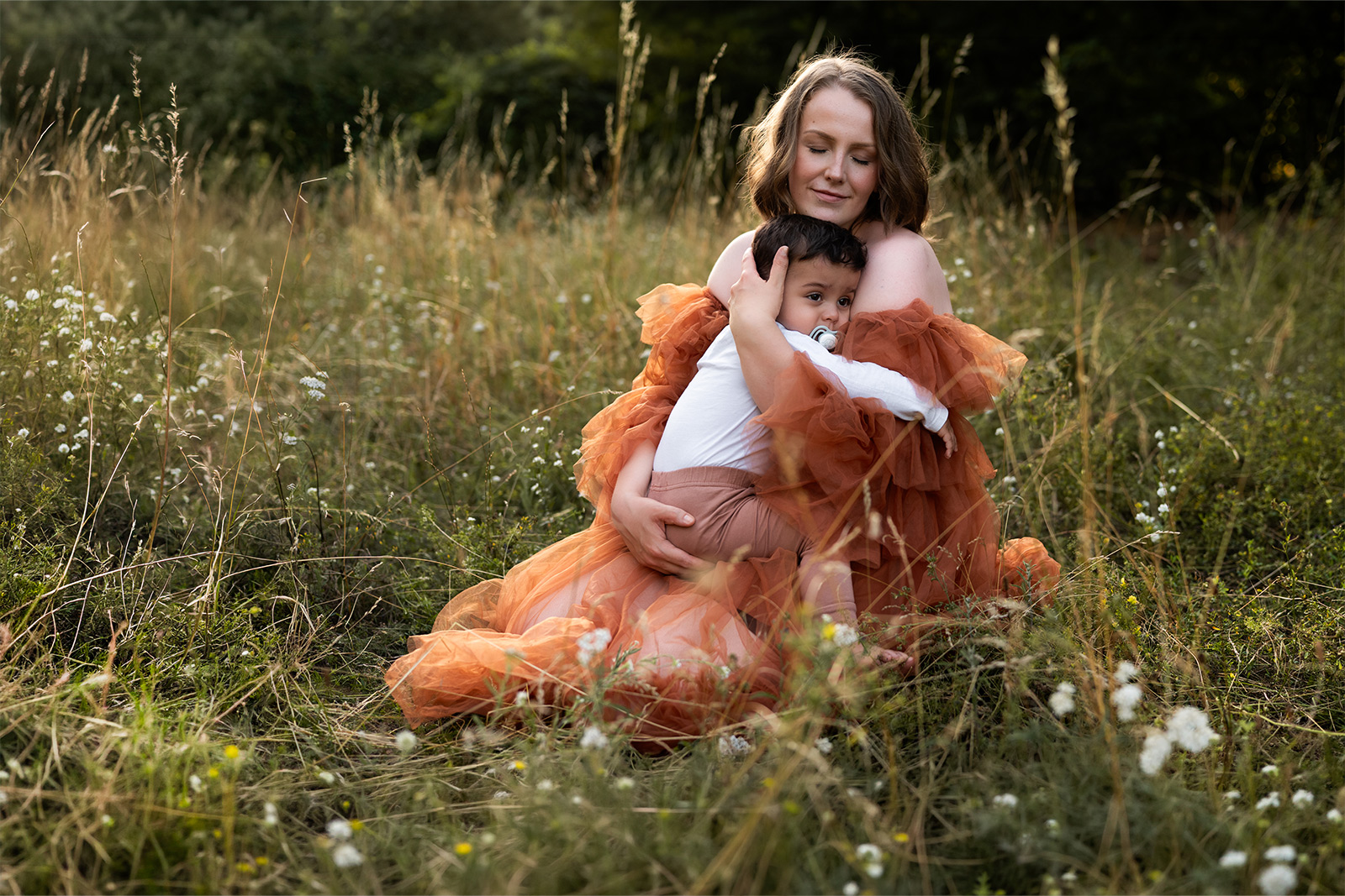 Eine Mutter im eleganten braunen Tüllkleid sitzt im hohen Gras und umarmt ihr kleines Kind liebevoll. Emotionale Familienfotografie im Freien in Deutschland.