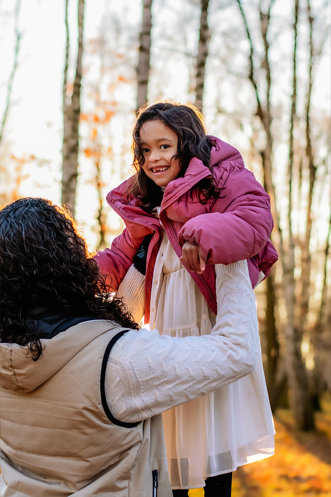 Eine Mutter hebt ihre lachende Tochter in einer rosa Jacke im sonnigen Wald hoch. Herzliche Kinder- und Familienfotografie in der Natur in Deutschland.