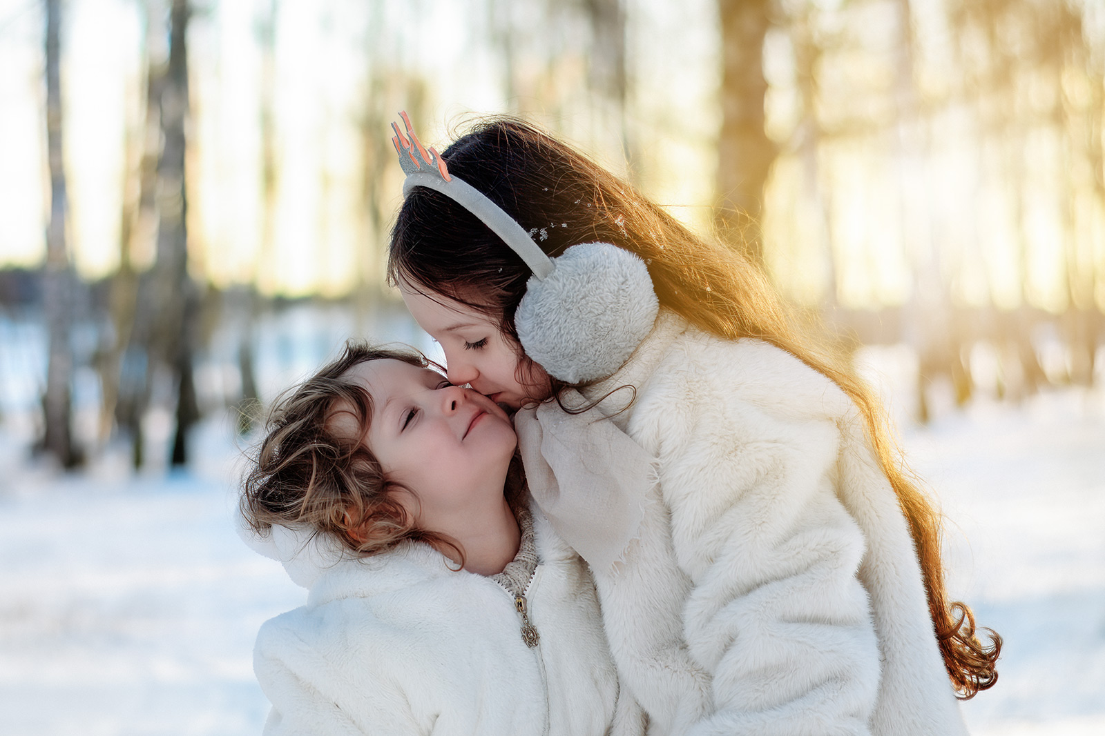 Ein älteres Mädchen küsst seine kleine Schwester liebevoll auf die Wange im verschneiten Wald. Emotionale Familienfotografie im Winter in Deutschland.