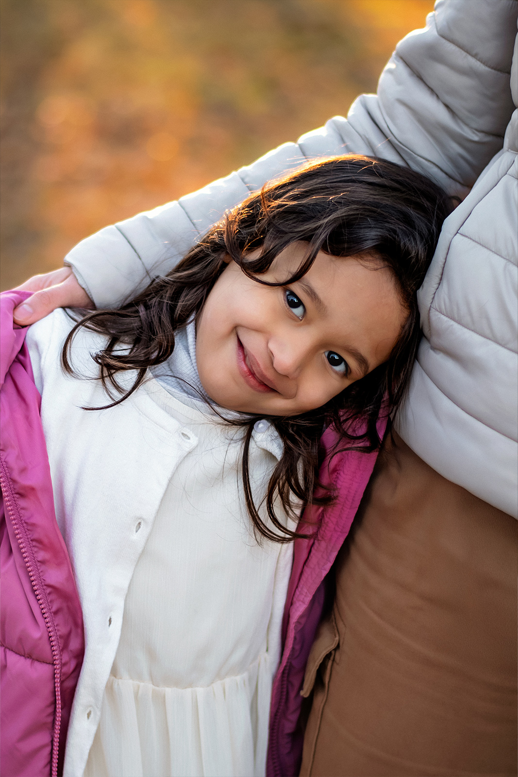 Ein kleines Mädchen mit dunklen Locken und rosa Jacke lehnt sich liebevoll an ihre Mutter bei einem Fotoshooting im Freien. Herzliche Familienfotografie in Deutschland.