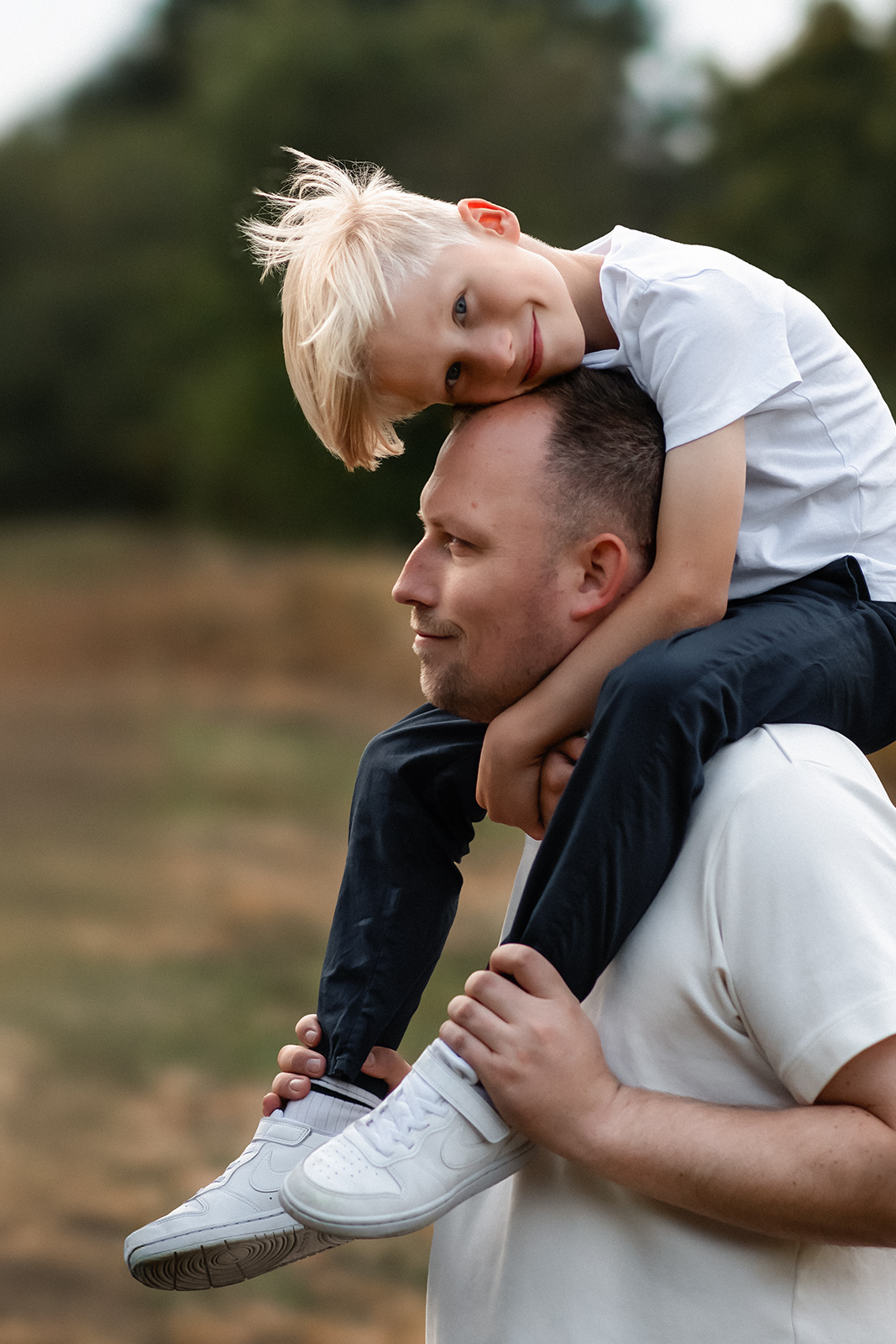 Ein blonder Junge sitzt auf den Schultern seines Vaters und stützt seinen Kopf liebevoll auf ihm ab bei einem Fotoshooting im Freien in Deutschland.