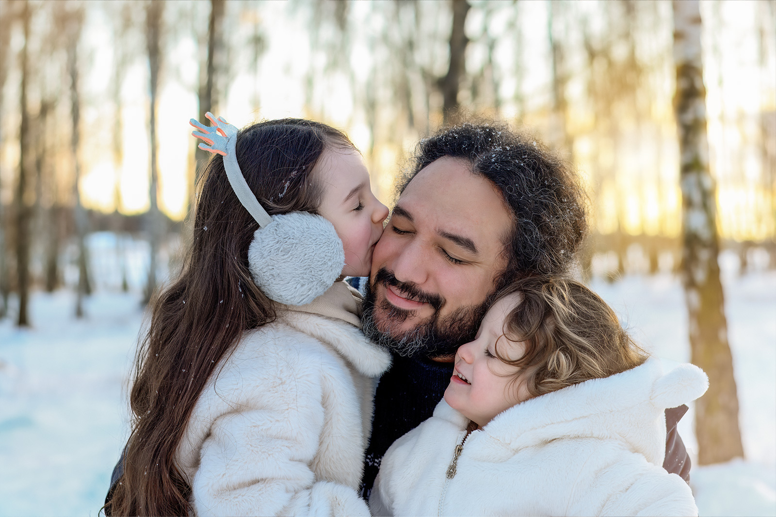 Ein glücklicher Vater umarmt seine zwei kleinen Töchter im verschneiten Wald, während eine Tochter ihn liebevoll auf die Wange küsst. Emotionale Familienfotografie im Winter in Deutschland.