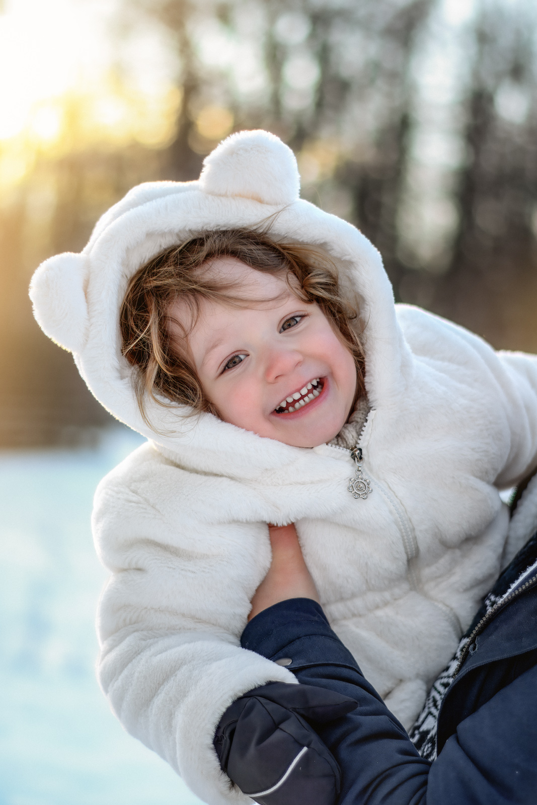 Ein fröhliches Kleinkind in einer weißen Winterjacke mit Kapuze und Bärenohren lächelt im verschneiten Freien. Süße Kinderfotografie im Winter in Deutschland.