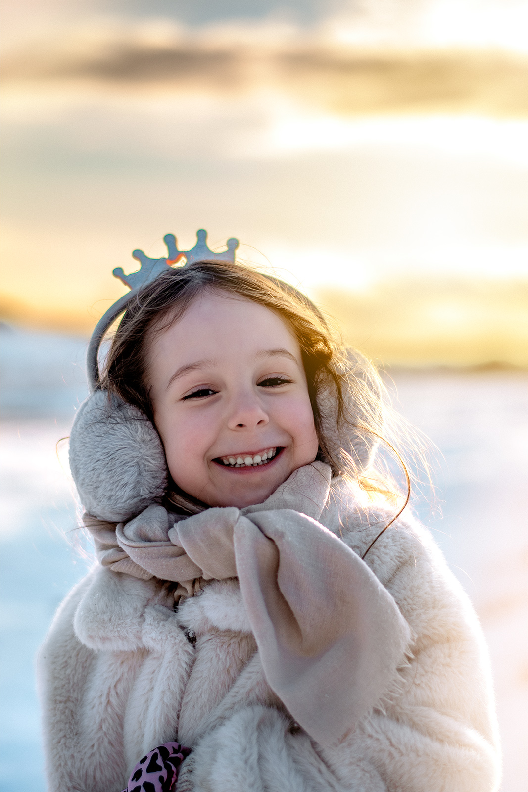 Ein strahlendes kleines Mädchen mit Ohrenschützern und Kronen-Haarreif lächelt glücklich im Schnee im warmen Sonnenlicht. Magische Kinderfotografie im Winter in Deutschland.