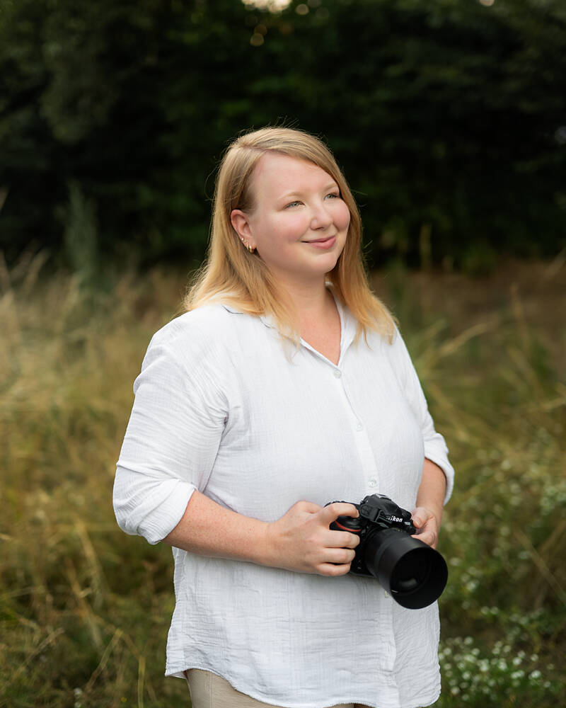 Berufliches Portr&auml;t der Fotografin Nicole de Melo, die mit einer DSLR-Kamera in der Hand in einem nat&uuml;rlichen Feld in Deutschland steht. Sie l&auml;chelt sanft und schaut zur Seite. Sie tr&auml;gt ein wei&szlig;es Leinenhemd. Nat&uuml;rliche Fotografie.