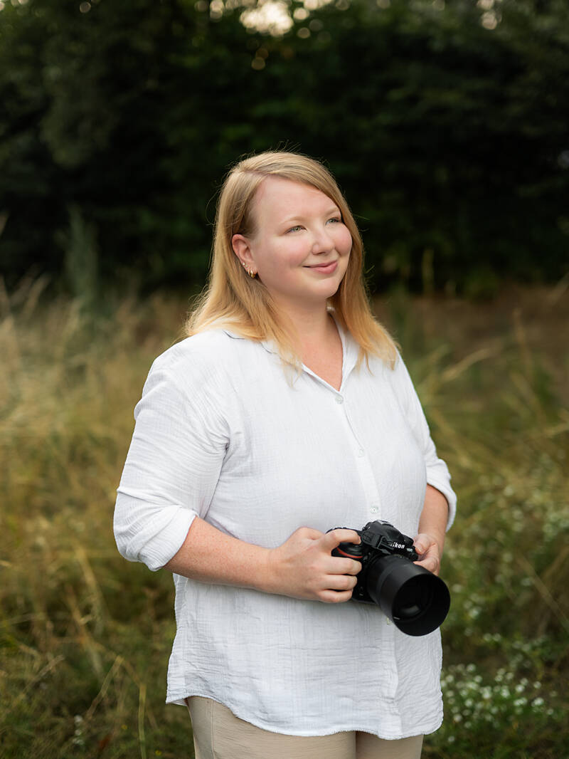 Berufliches Portr&auml;t der Fotografin Nicole de Melo, die mit einer DSLR-Kamera in der Hand in einem nat&uuml;rlichen Feld in Deutschland steht. Sie l&auml;chelt sanft und schaut zur Seite. Sie tr&auml;gt ein wei&szlig;es Leinenhemd. Nat&uuml;rliche Fotografie.