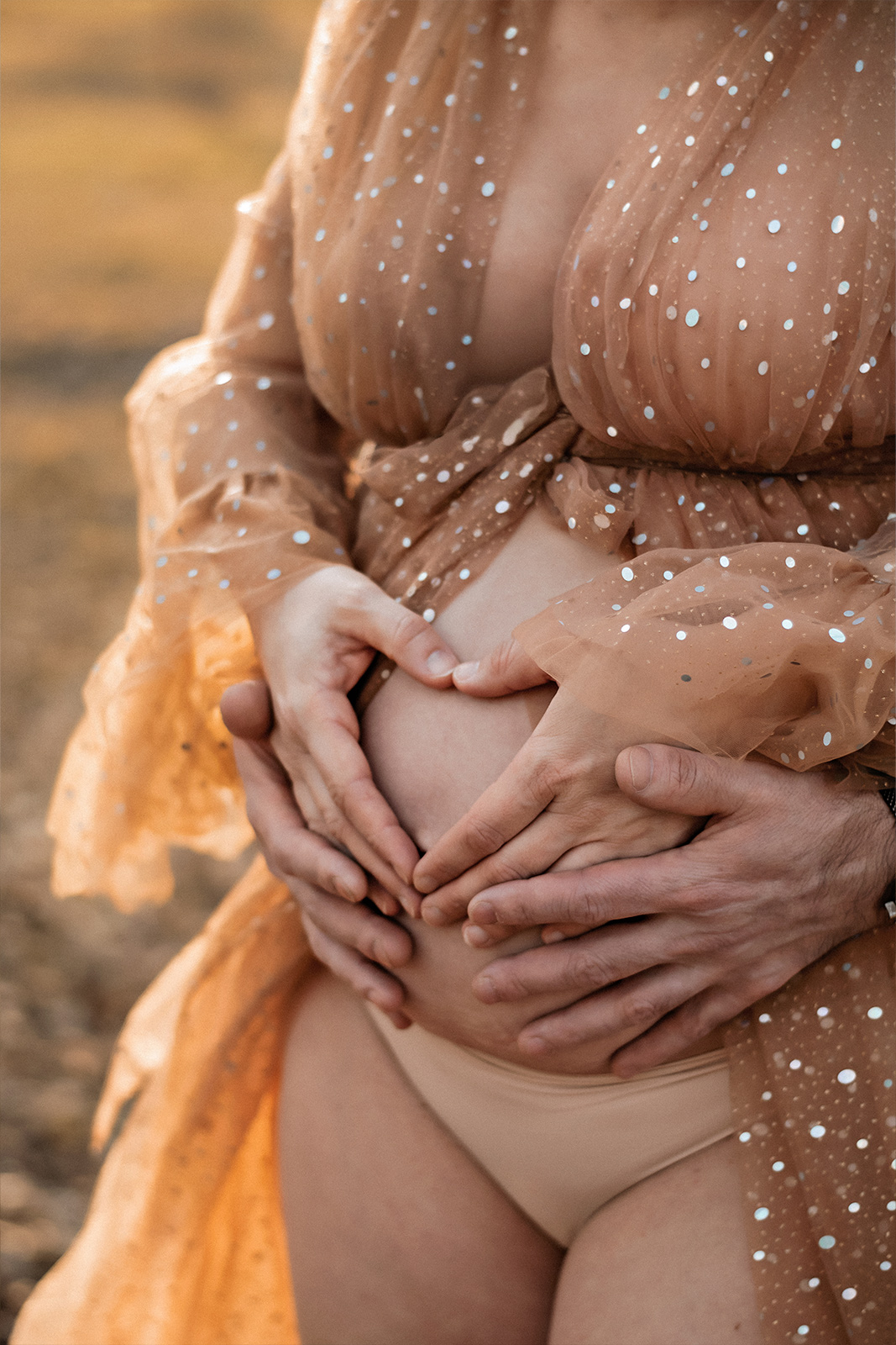 Nahaufnahme eines Babybauchs in einem glitter Tüllkleid, umrahmt von den Händen der Eltern, die ein Herz formen. Detailreiche Schwangerschaftsfotografie in Deutschland.