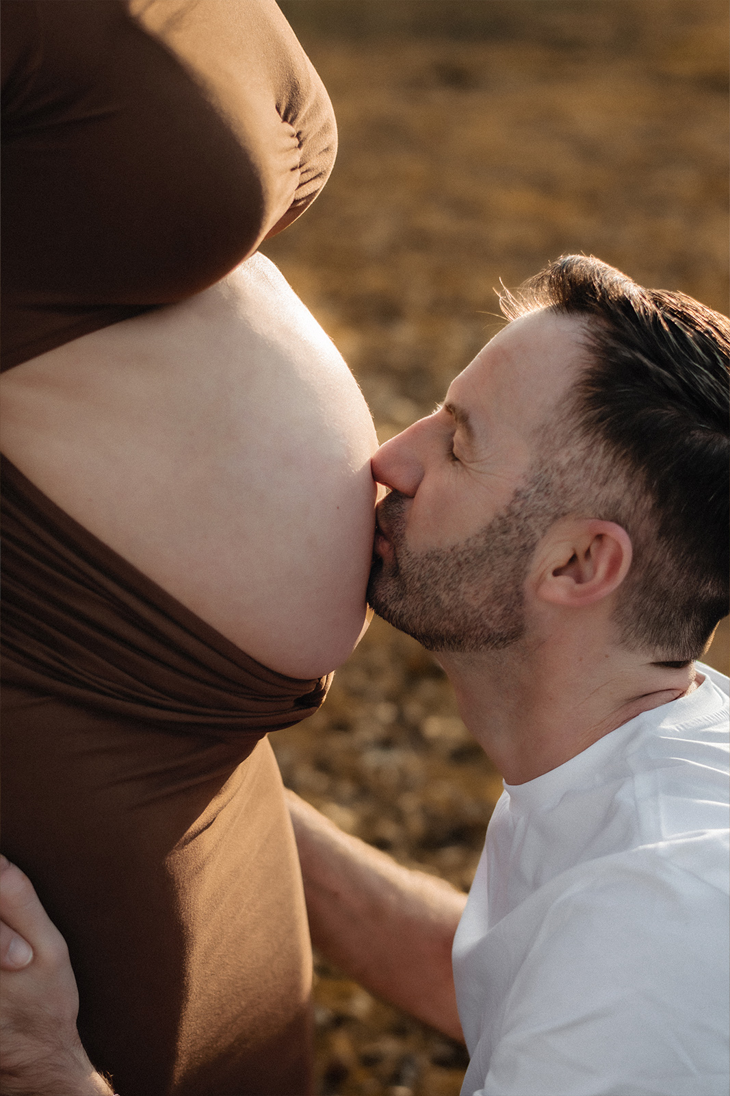 Nahaufnahme eines werdenden Vaters, der liebevoll den Babybauch seiner Frau küsst. Emotionale Schwangerschaftsfotografie im Freien in Deutschland.