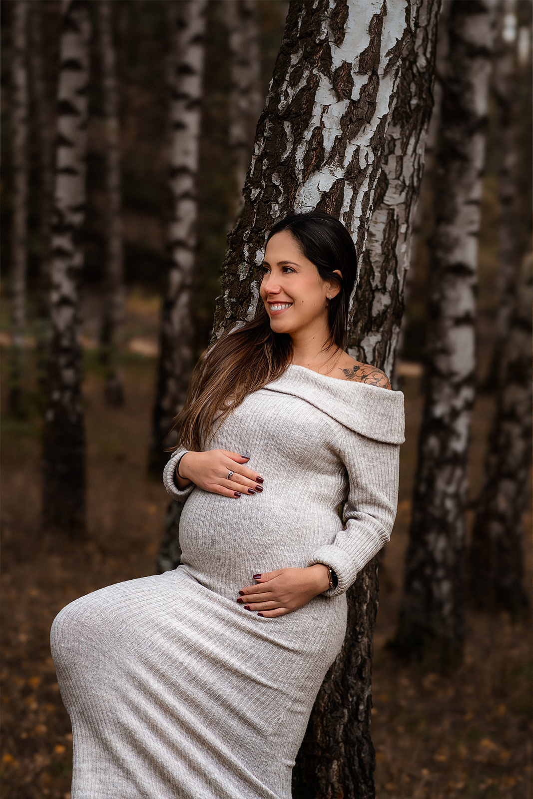 Eine lächelnde schwangere Frau im grauen Strickkleid lehnt an einem Birkenstamm im Wald. Ästhetische Babybauch Fotografie im Freien in Deutschland.