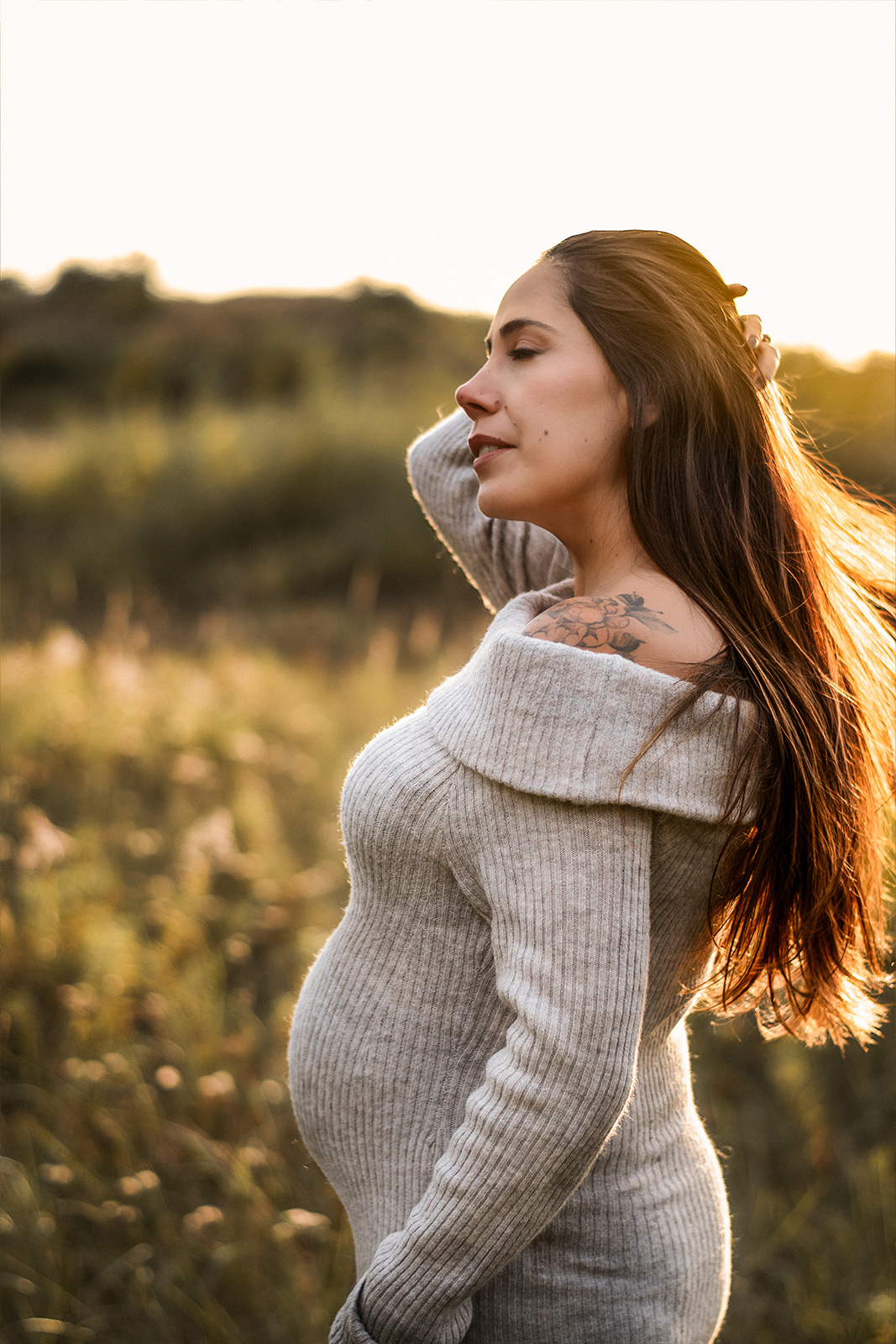 Eine schwangere Frau im beigen Strickkleid steht bei Sonnenuntergang in hohem Gras auf einer Wiese. Ästhetische Babybauch Fotografie im Freien in Deutschland.