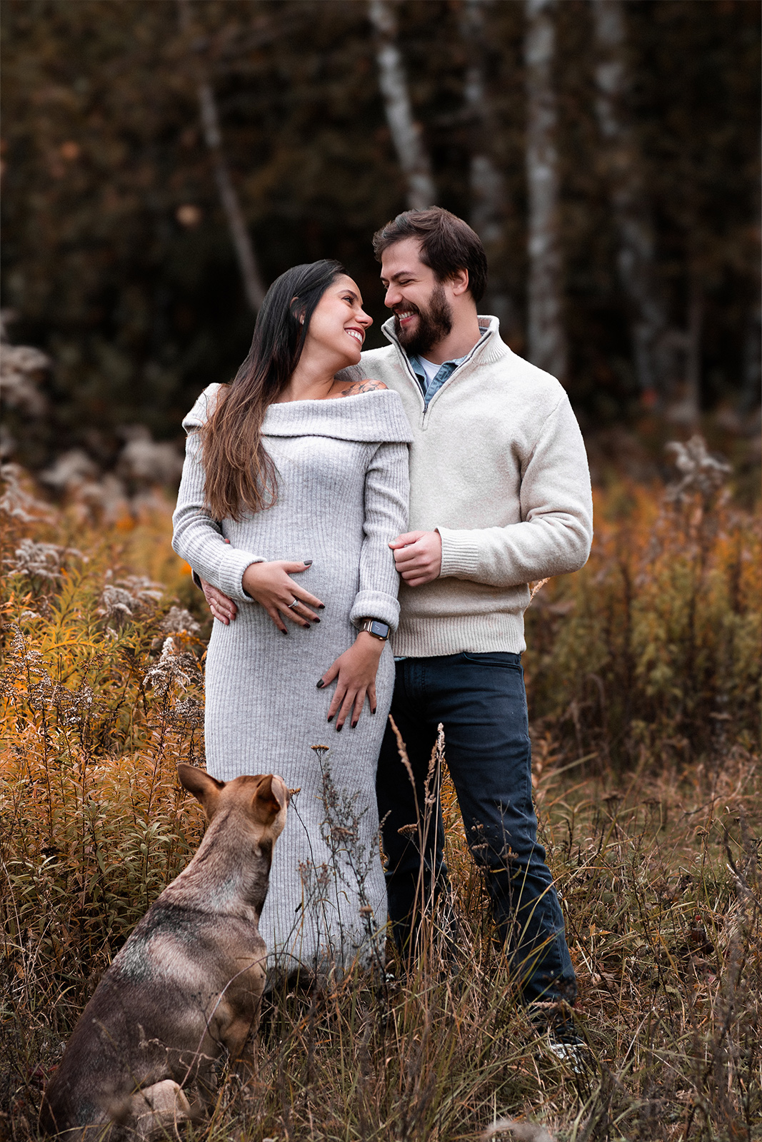Ein werdendes Elternpaar steht mit seinem Hund auf einer Herbstwiese bei einem Babybauch Shooting im Freien in Deutschland.