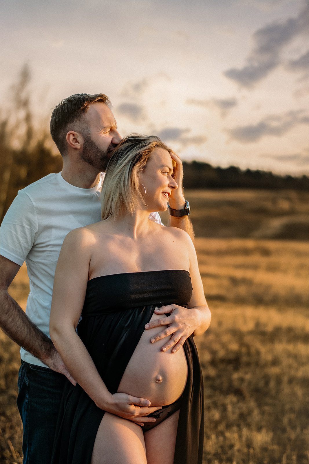 Ein werdender Vater küsst seine Frau im schwarzen Kleid liebevoll auf den Kopf während eines Babybauch Shootings im Freien bei Sonnenuntergang in Deutschland.