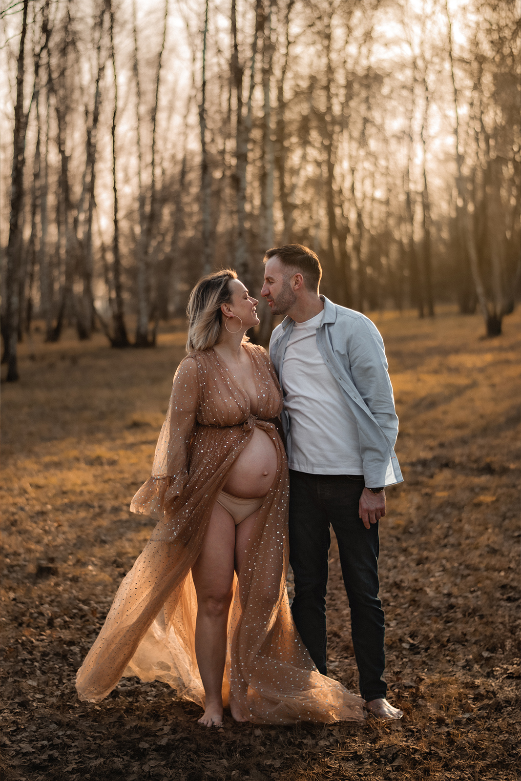 Ein werdendes Elternpaar steht zusammen vor einer Kulisse aus Birkenbäumen. Die Frau trägt ein fließendes Glitzer-Tüllkleid bei einem Babybauch Shooting im Freien in Deutschland.