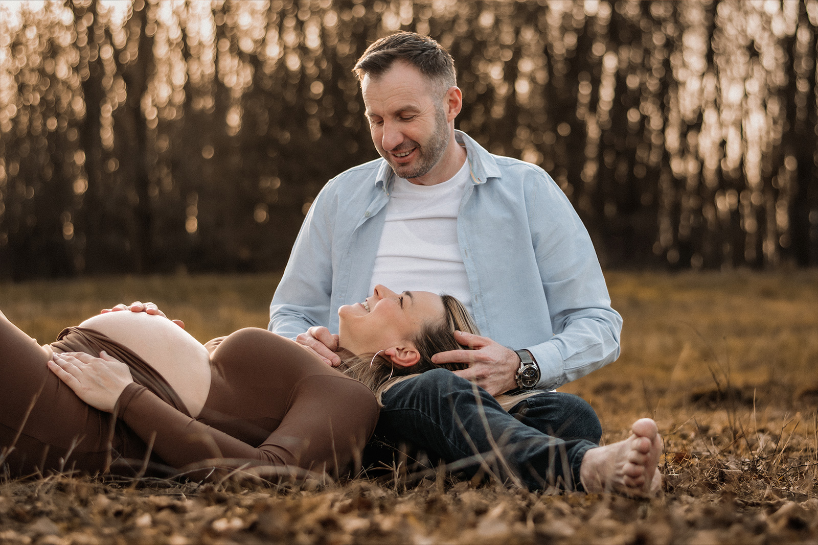 Glückliche schwangere Frau liegt im Schoß ihres Mannes auf einer Wiese im Freien bei einem Babybauch Shooting in Deutschland.